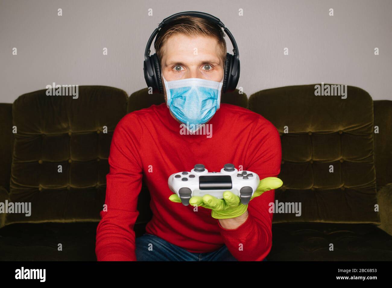 Mann, der Videospiele spielt, trägt eine chirurgische Maske, Gummihandschuhe und Kopfhörer mit Geräuschminimierung. Selbstisolierung (Selbstquarantäne) für Coronavirus (COVID Stockfoto