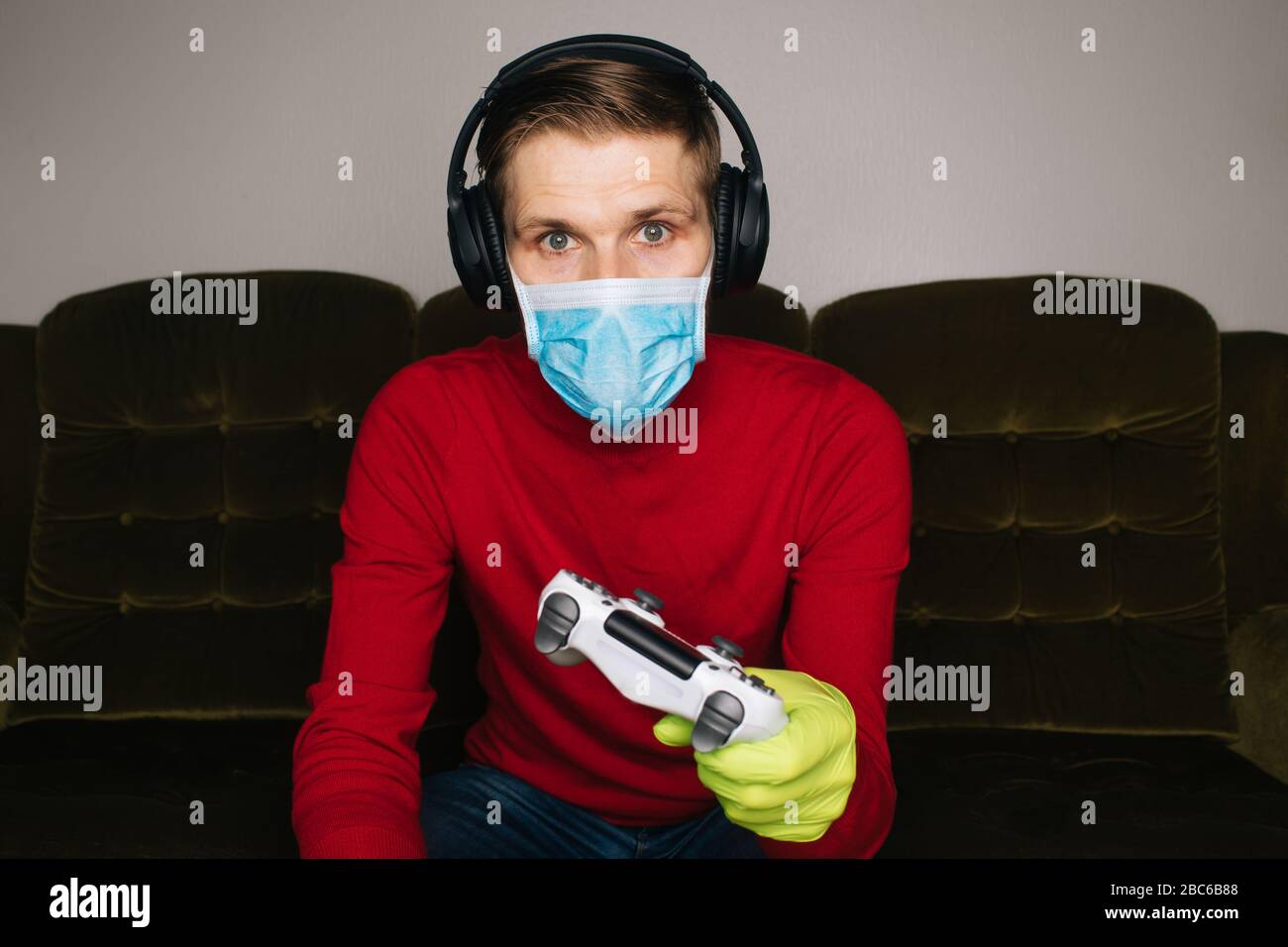 Mann, der Videospiele spielt, trägt eine chirurgische Maske, Gummihandschuhe und Kopfhörer mit Geräuschminimierung. Selbstisolierung (Selbstquarantäne) für Coronavirus (COVID Stockfoto