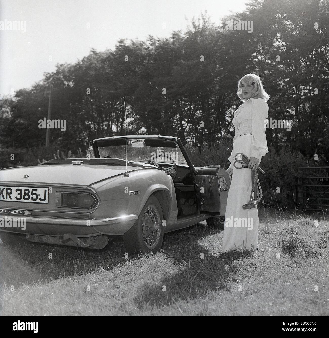 1970er Jahre, historisch, draußen auf einem sonnenbeleuchteten Feld, eine attraktive blonde Frau in einer weißen Oberteil- und glockenuntergebrachten Hose, neben ihrem offenen Sportwagen, einem Triumph Spitfire IV, mit Drahträdern, England, Großbritannien stehend. Der 1962 erstmals vorgestellte Spitfire, der auf einem Entwurf von Giovanni Michelotti basierte, war ein kleiner britischer Zweisitzer-Sportwagen und wurde bis 1980 hergestellt. Das hier abgebildete Mark IV-Modell wurde in den Jahren 1970 bis 1974 bei Standard-Triumph Works (im Besitz von Leyland Motors) in Canley, Coventry hergestellt und neben anderen neuen Merkmalen aus früheren Modellen war ein neu gestaltetes Heckende Stockfoto