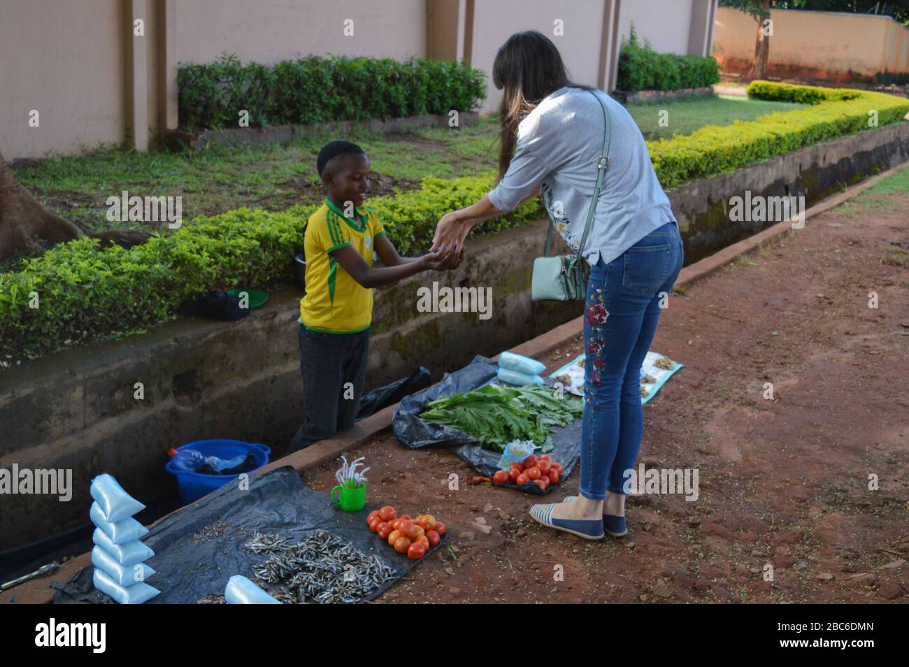 LILONGWE, MALAWI, AFRIKA - 2. APRIL 2018: Ein afrikanischer malawischer kleiner Junge verkauft Tomatos, Fisch und grüne Blätter entlang der Straße. Junge nimmt Kandi Stockfoto