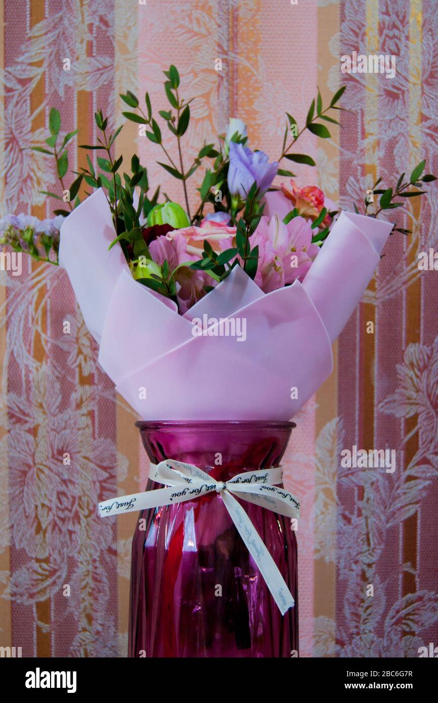 Ein zartes Blumenstrauß verschiedener Blumen in einem hellrosa Wrapper steht vor einem Hintergrund rosafarbener Tapeten in einer Hauseinstellung. Vertikales Foto. Stockfoto