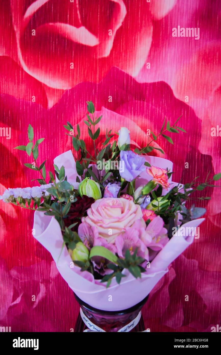 Ein zartes Blumenstrauß mit verschiedenen Blumen in einem hellrosa Wrapper steht in einer häuslichen Umgebung gegen eine bunte rosafarbene Wand. Vertikales Foto. Stockfoto