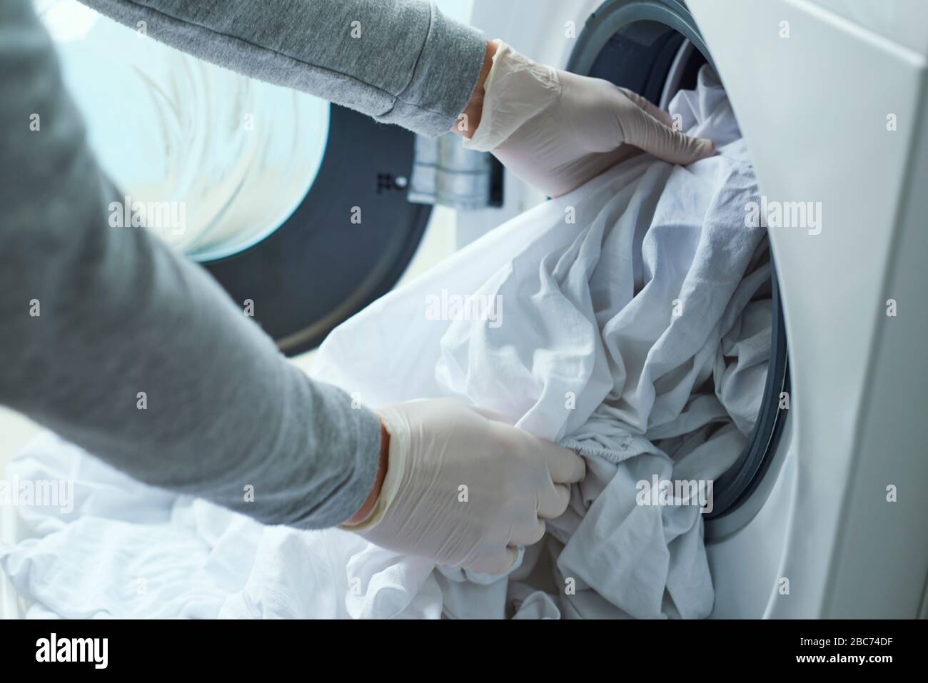 Nahaufnahme eines jungen kaukasischen Mannes, der Latexhandschuhe trägt und weiße Bettwäsche in die Waschmaschine einbringt Stockfoto