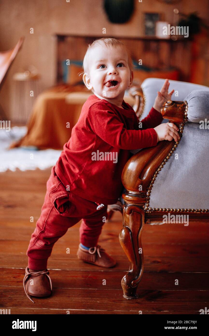 Ein kleiner niedlicher Junge in einer roten Jacke und Hose, neben einem weichen Stuhl stehend Stockfoto