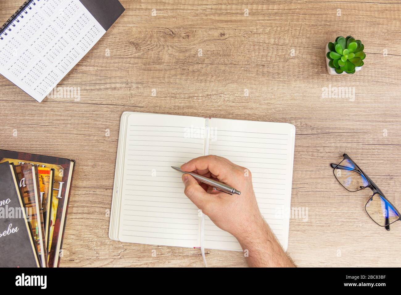 Männliche Hände schreiben in einem Notizbuch am Büroschreibtisch mit Glas, Büchern, sükulent auf Holzhintergrund Stockfoto
