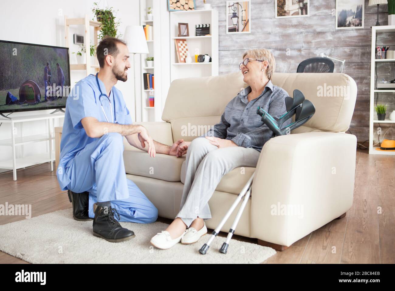 Freudige alte Frau in einem Pflegeheim, die mit ihrem Gesundheitsbesucher spricht, während sie mit Krücken neben ihr auf der Couch sitzt. Stockfoto