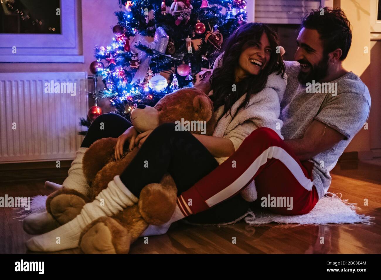 Ein Paar lacht und umarmt vor dem Weihnachtsbaum. Mädchen hält Teddybär Stockfoto