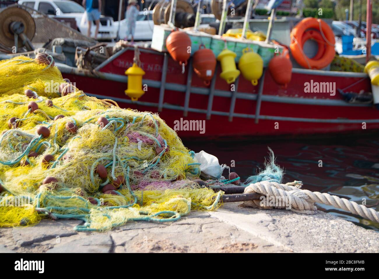 An der Seite eines Yachthafens auf der griechischen Insel Kreta stapelten sich Fischernetze, im Hintergrund ein kleines rotes Fischerboot Stockfoto