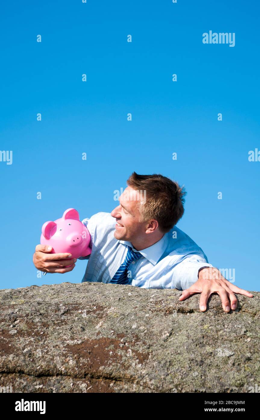 Lächelnd Geschäftsmann halten auf einen treuen rosa Sparschwein, wie er oben auf einem felsigen Klippe erreicht. Stockfoto