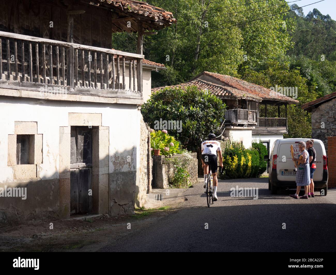 Straßenradsport in Asturien, Nordspanien. Rennradrennfahrerin geht an zwei Frauen vorbei, die Brot von einer mobilen Bäckerei im Dorf Riocaliente im Pico kaufen Stockfoto