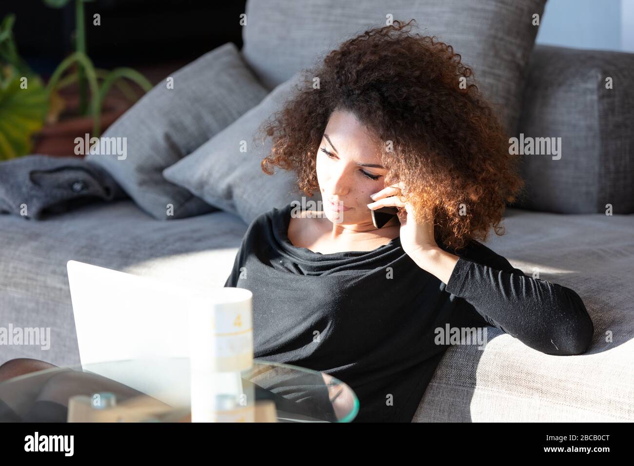 Junge Frau, die während des Lockdowns für die Covid-19-Pandemie von zu Hause aus arbeitet und sich mit ihrem Laptop und Handy in der Sonne auf einem Sofa entspannt Stockfoto