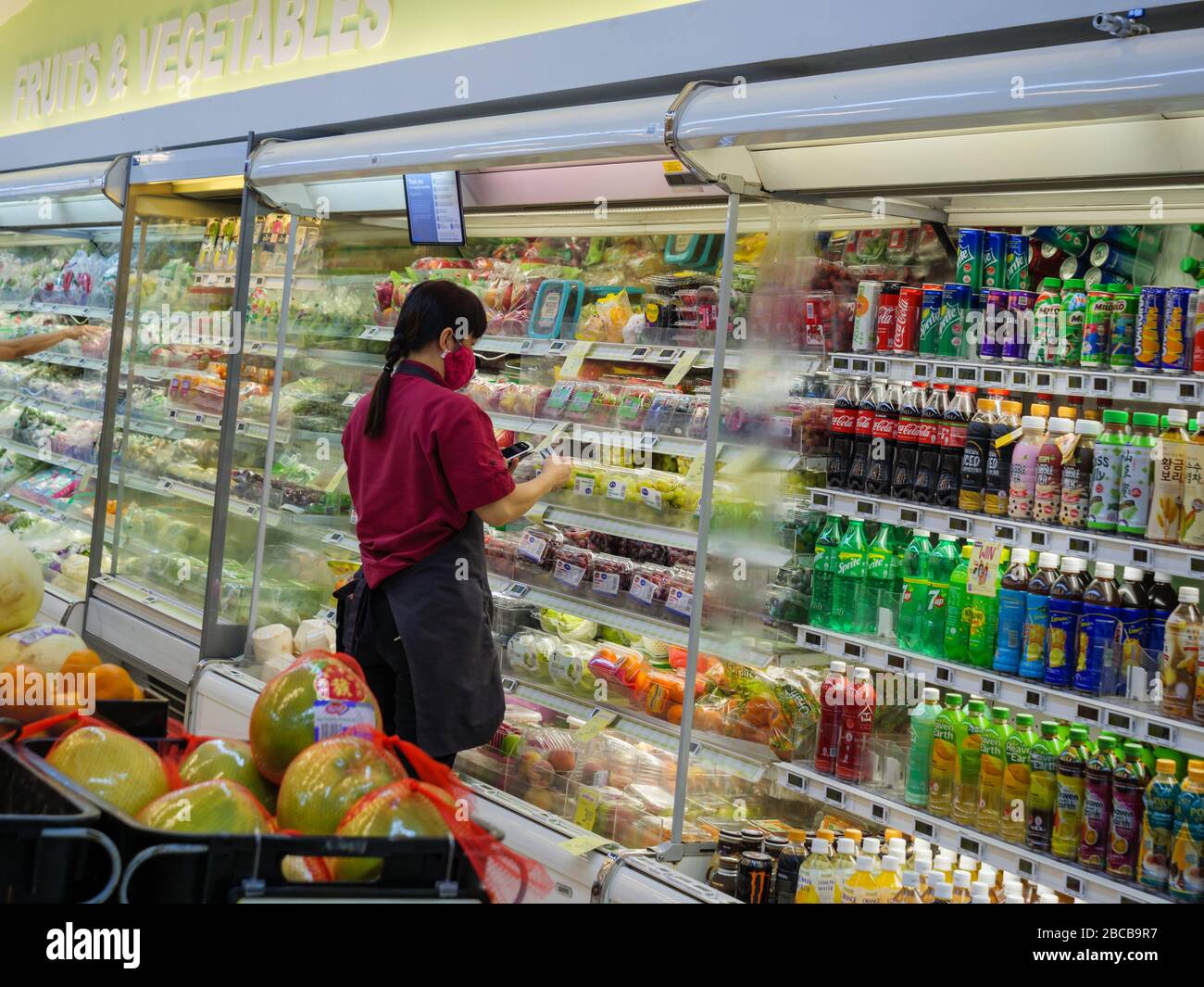 SINGAPUR - 2. APR 2020 - maskenbekleideten Supermarktmitarbeiter kleben Etiketten auf Obst und Gemüse in einem NTUC FairPrice Supermarkt Stockfoto