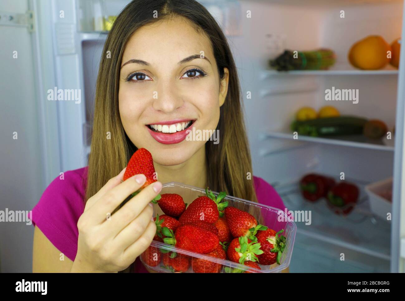 Nahaufnahme des hübschen lächelnden Mädchens in der Nähe des Kühlschranks, der Erdbeeren in den Händen hält, blickt auf die Kamera. Gesundes Lebensmittelkonzept. Frisches Obst und Gemüse Stockfoto