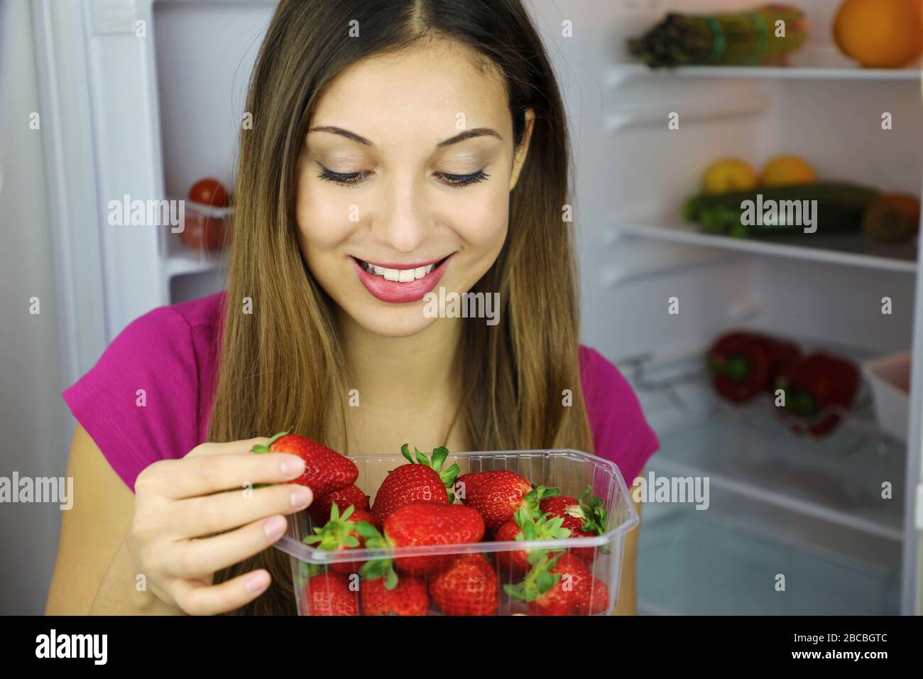 Nahaufnahme des hübschen lächelnden Mädchens in der Nähe des Kühlschranks und Blick auf Erdbeeren. Gesundes Lebensmittelkonzept. Frisches Obst und Gemüse. Stockfoto