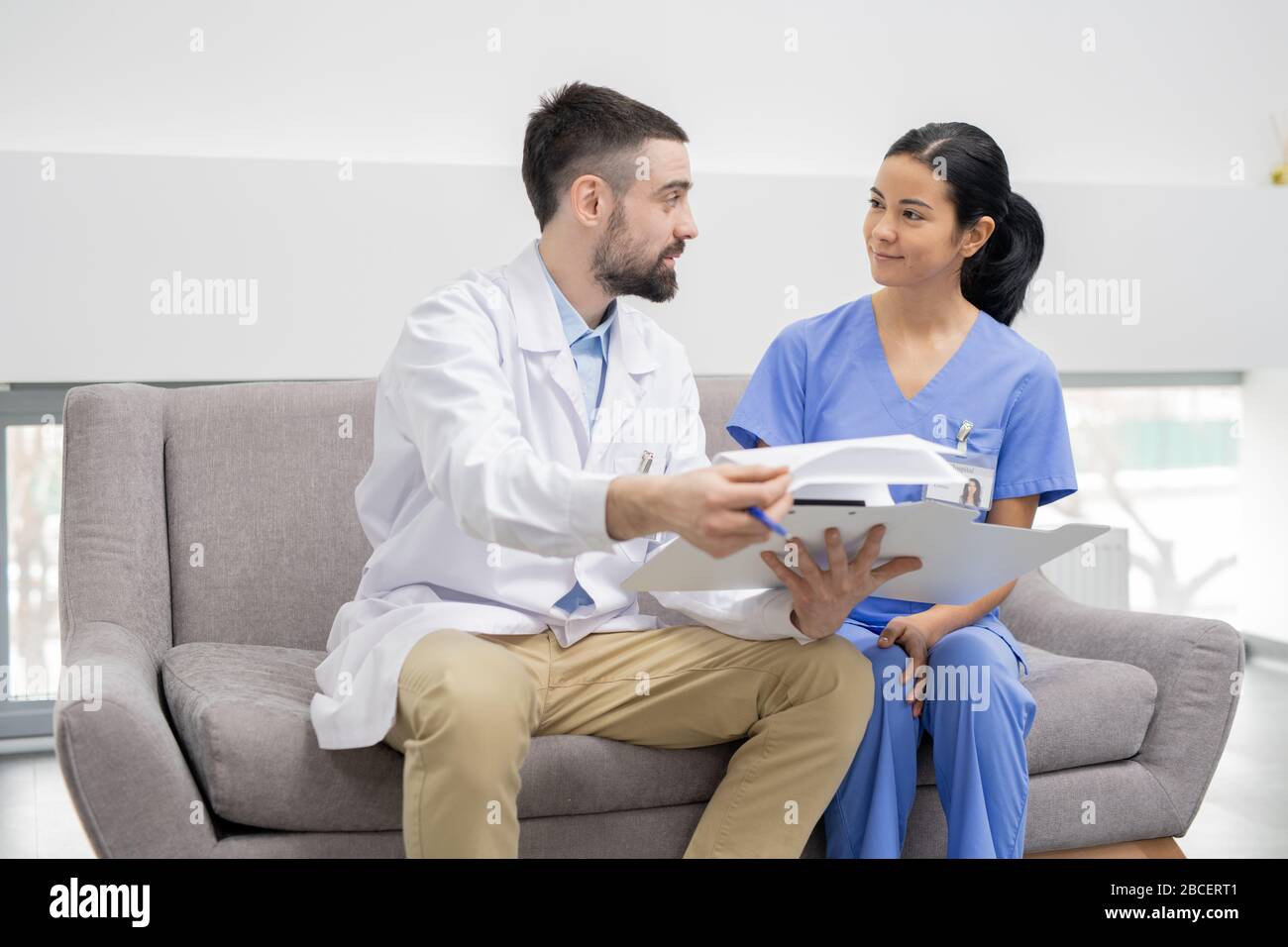 Der junge Arzt in Whitecoat erklärt seinem hübschen Assistenten in blauer Uniform die Informationen in einem Dokument, während er in der Lobby der Kliniken sitzt Stockfoto