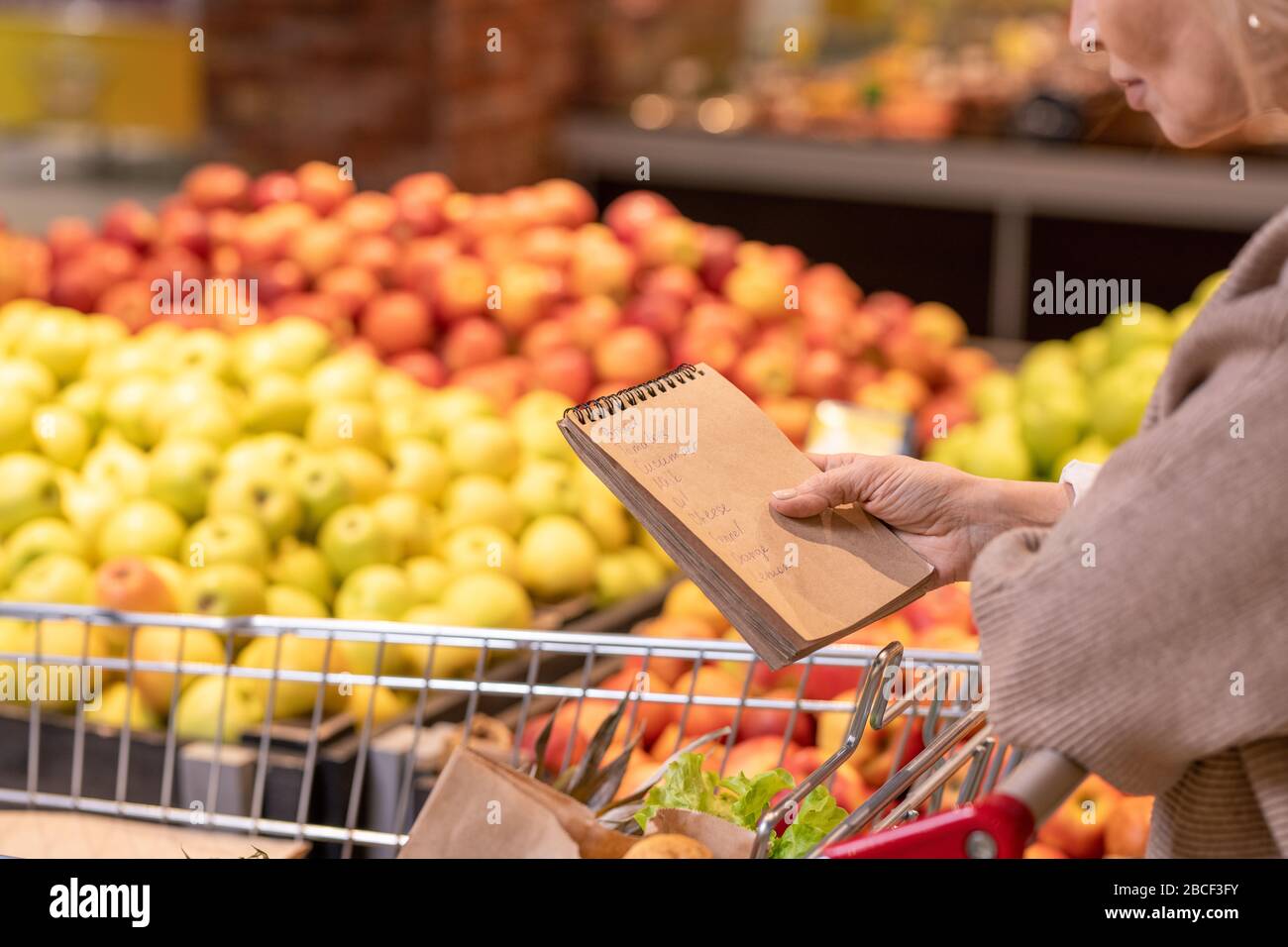 Reifer weiblicher Konsument mit Notizblock über Lebensmittelprodukten im Einkaufswagen, der durch die Einkaufsliste schaut, während er sich auf der Obstanzeige bewegt Stockfoto