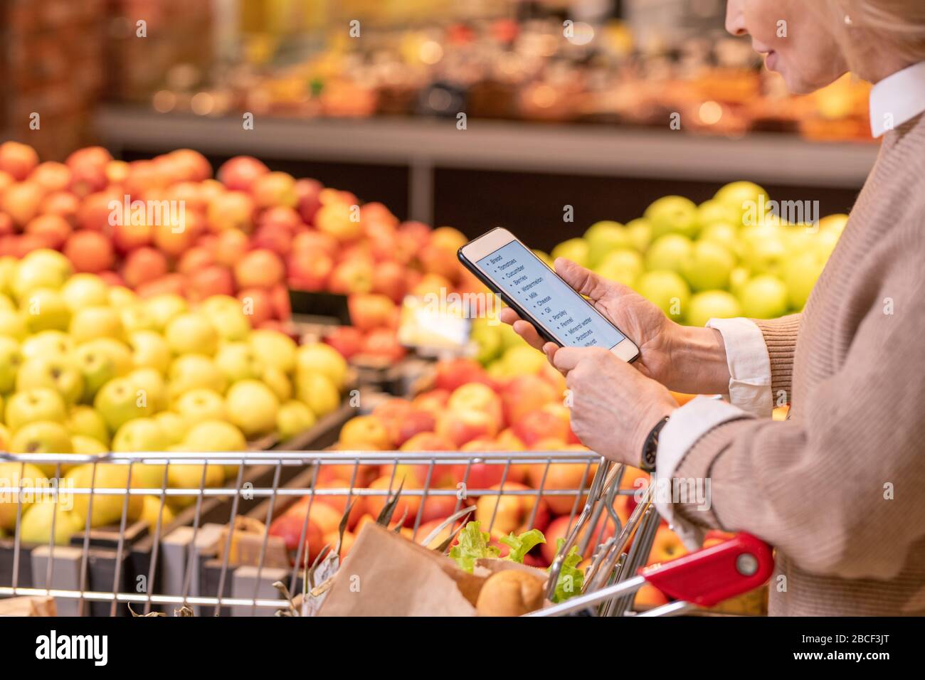 Moderne, reife weibliche Kundin scrollt durch die Liste der Lebensmittelprodukte, die im Supermarkt gekauft werden sollen, während sie an der Fruchtanzeige vorbeigeht Stockfoto