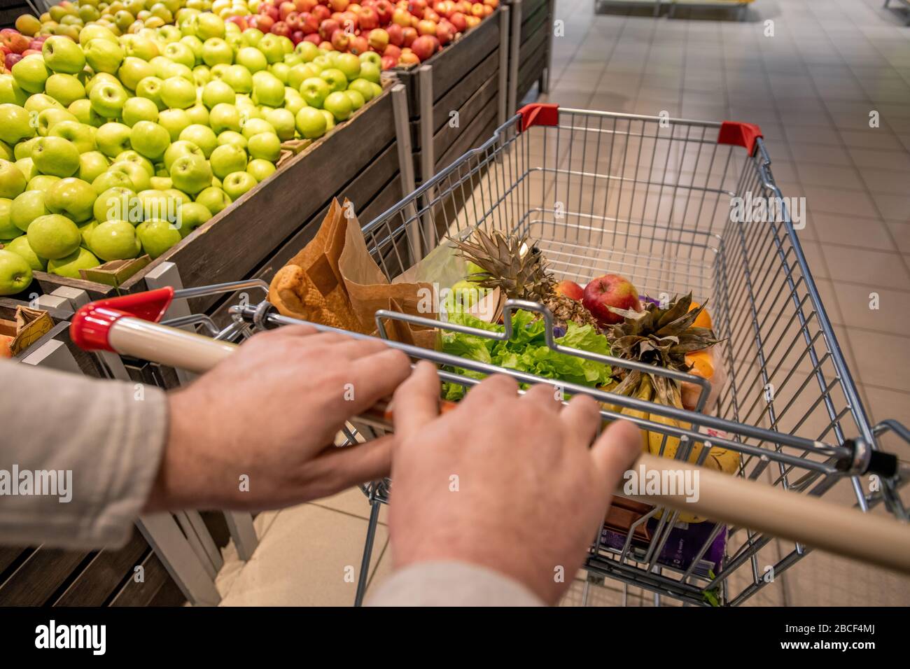 Hände von alten männlichen Kunden, die den Einkaufswagen mit Lebensmitteln bedrängen, während sie mit frischen Äpfeln im Supermarkt auf dem Obsttäfelchen unterwegs sind Stockfoto