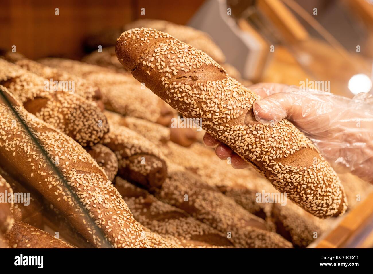 Handschuh von reifen weiblichen Kunden, die frisches Brot mit Sesamstreuen beim Kauf von Lebensmittelprodukten im modernen Supermarkt nehmen Stockfoto
