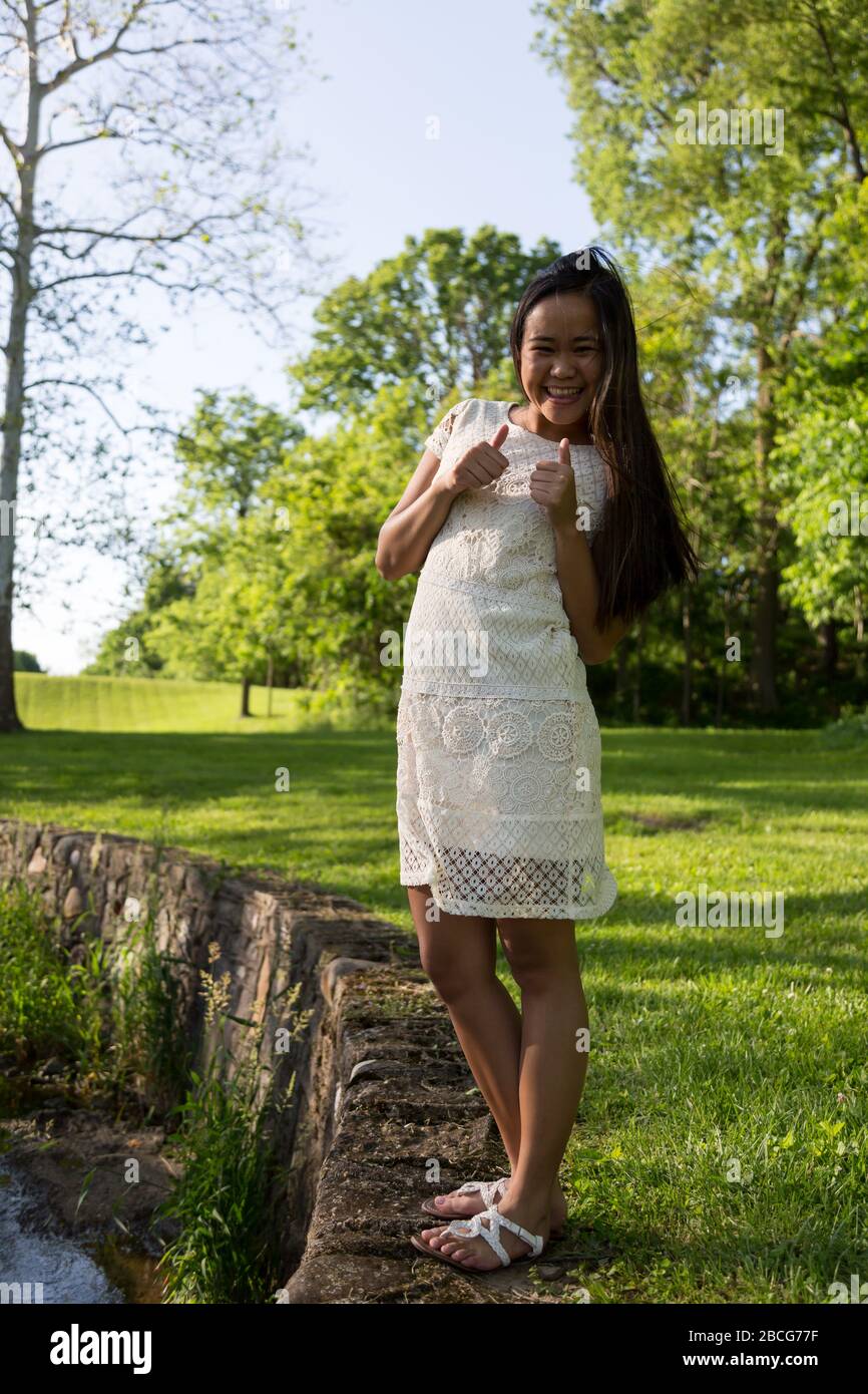 Ein schönes junges Filipina-Mädchen in Weiß gibt das Daumen-up-Zeichen, während sie für ihre Senior Pictures in Sunken Gardens in Huntington, Indiana, USA posiert. Stockfoto