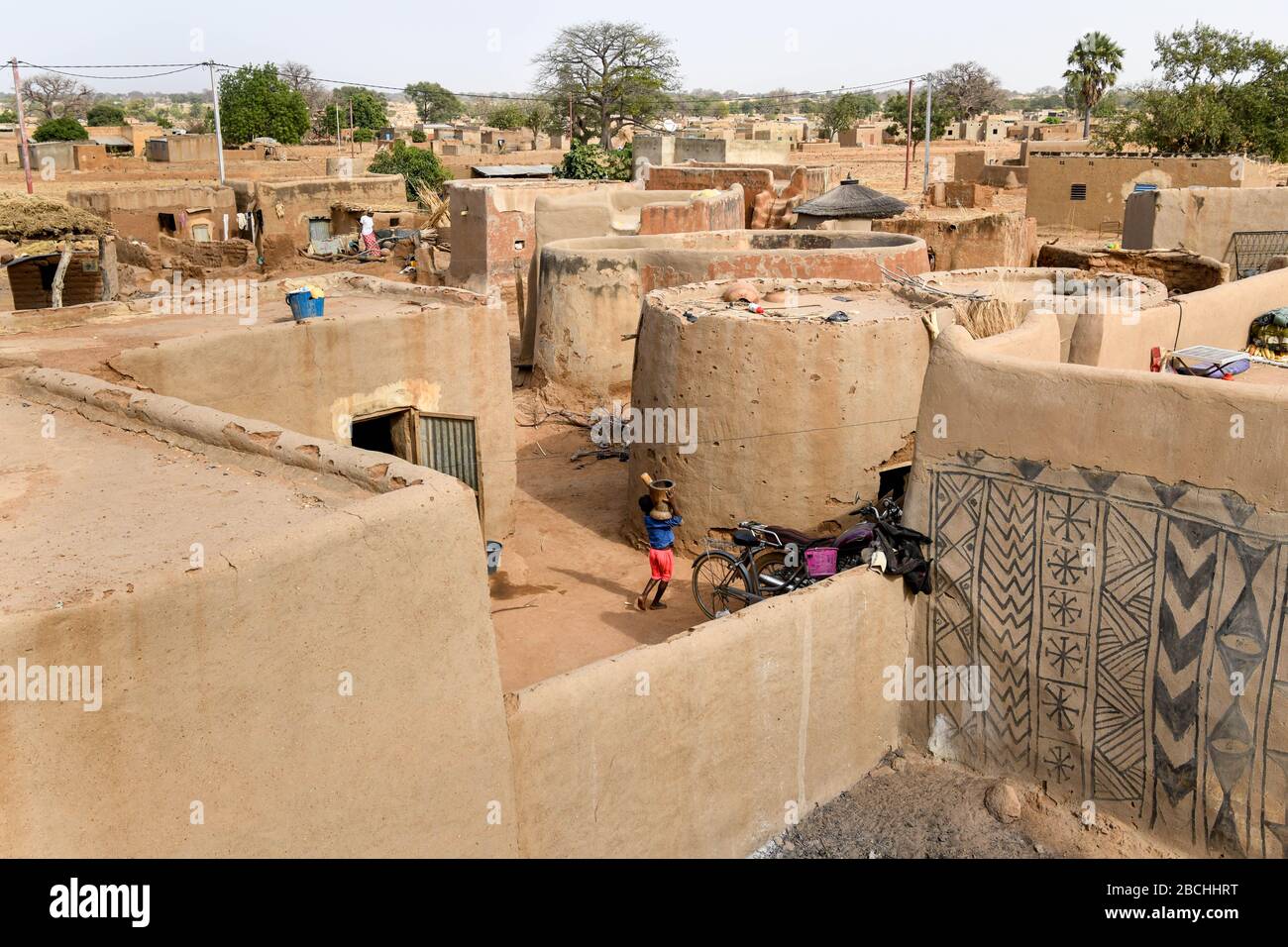 Afrika, Burkina Faso, Region Pô, Tiebele. Stadtansicht des königlichen Hofdorfes in Tiebele. Ein Kind geht mit einem traditionellen Grinder auf seinem s Stockfoto