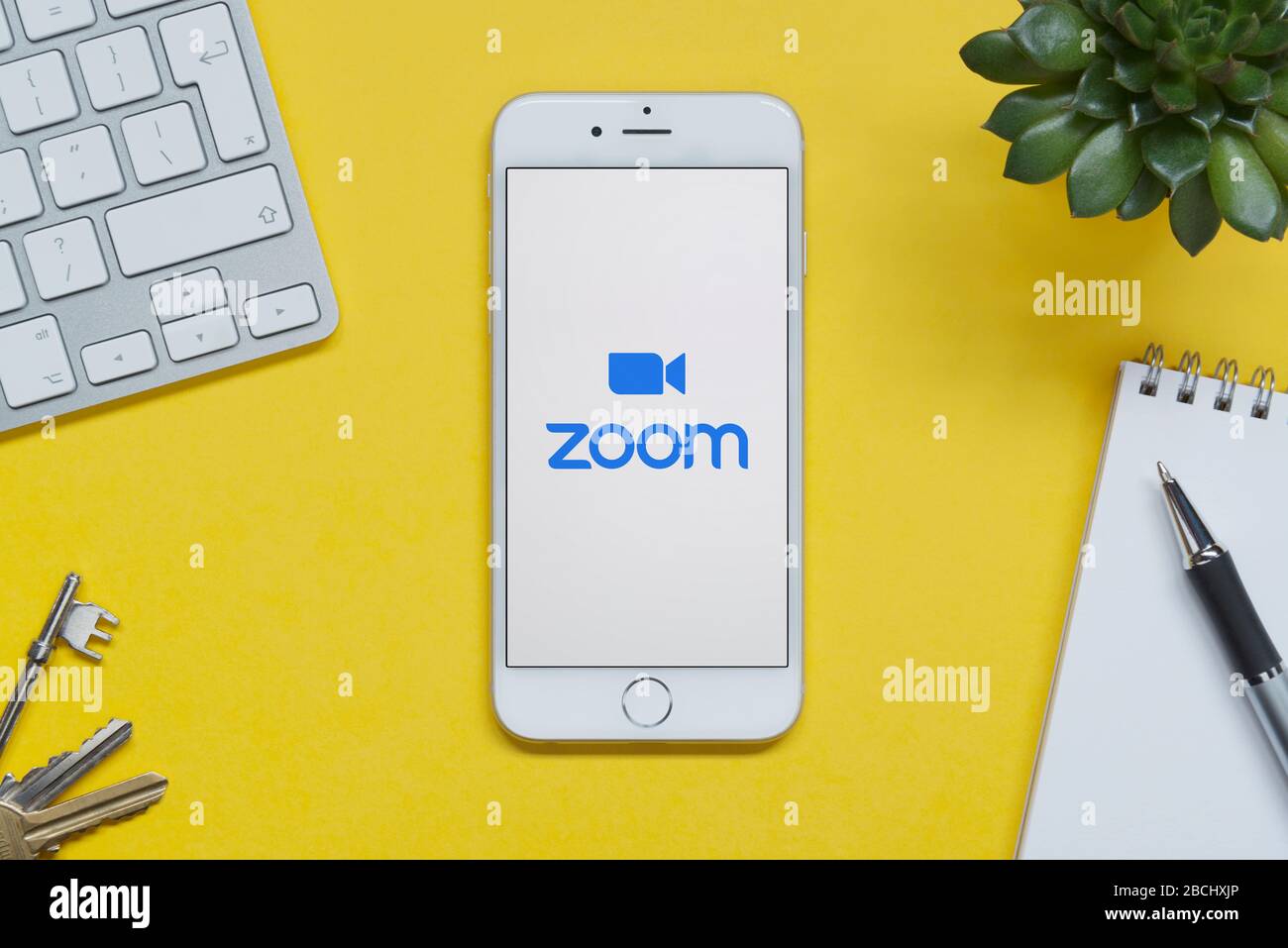 Ein iPhone mit Zoom-Logo liegt auf einem gelben Hintergrund Tisch mit Tastatur, Tasten, Notizblock und Pflanze (nur für redaktionelle Verwendung). Stockfoto