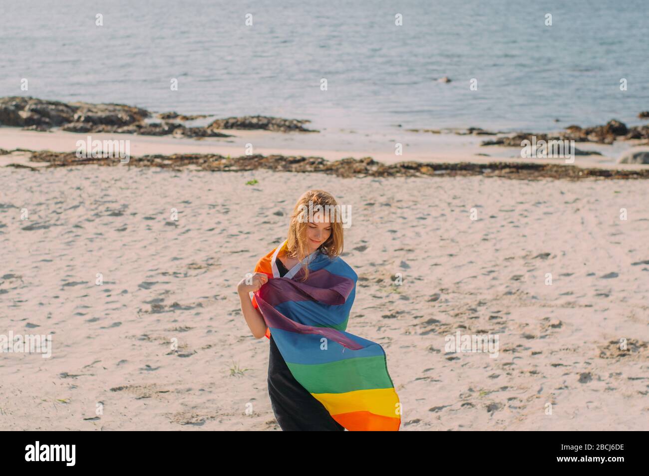 Junges blondes Mädchen mit LGBTQI-Flagge, das am Tag am weißen Strand in der Nähe des Meeres, Lofoten Islands, Norwegen spazieren ging Stockfoto