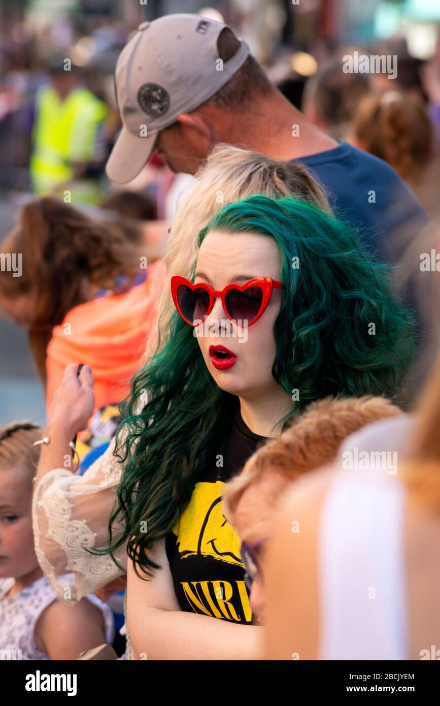 Extravagante junge Frau mit grünem Haar und roter Brille, die bei der Straßenparade einen lustigen Gesichtsausdruck macht Stockfoto
