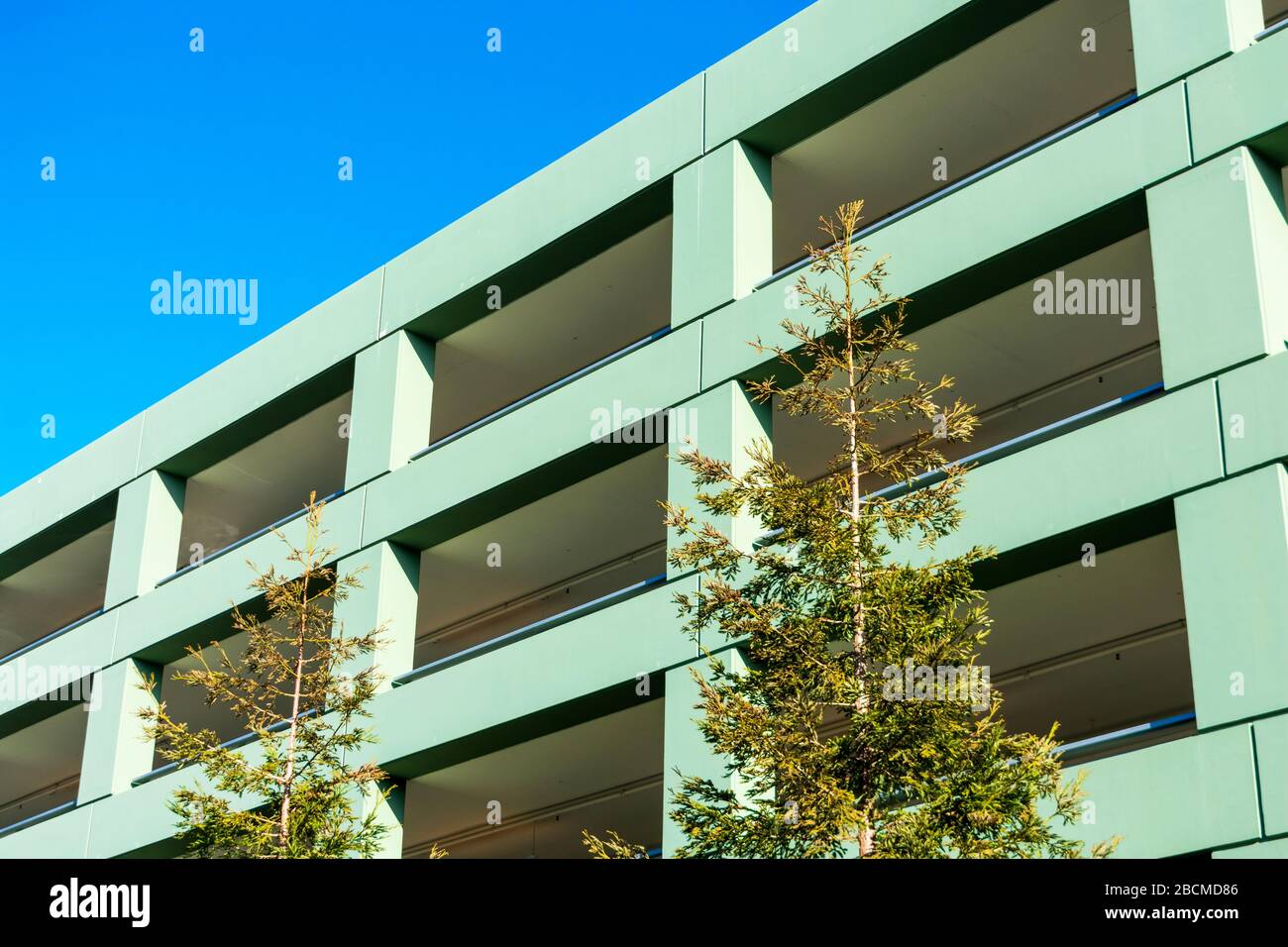 Parkhaus auf mehreren Ebenen, Fassade und Außenansicht. Grüne Vordergrundbäume. Blauer Himmel im Hintergrund. Stockfoto