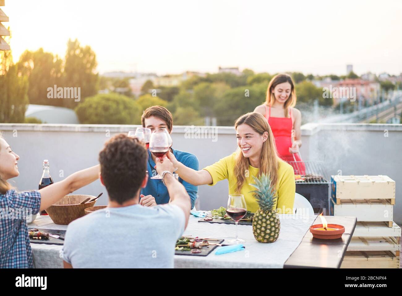 Junge Freunde, die bei Sonnenuntergang auf der Penthouse-Terrasse Grillparty haben - glückliche Leute, die ein grillabendessen im Freien machen, Fleisch kochen und Wein trinken - konzentrieren sich auf die Bohrinsel Stockfoto