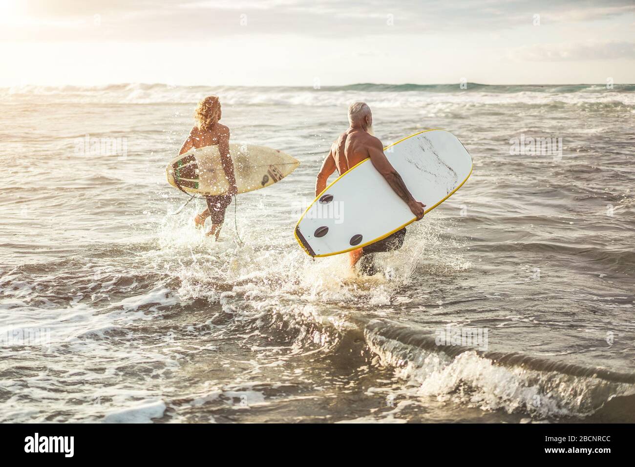 Verschiedene Altersfreunde, die bei Sonnenuntergang am Strand zum Surftraining laufen - Familienmitglieder haben Spaß beim Extremsport - fröhliche ältere und gesunde LIF Stockfoto