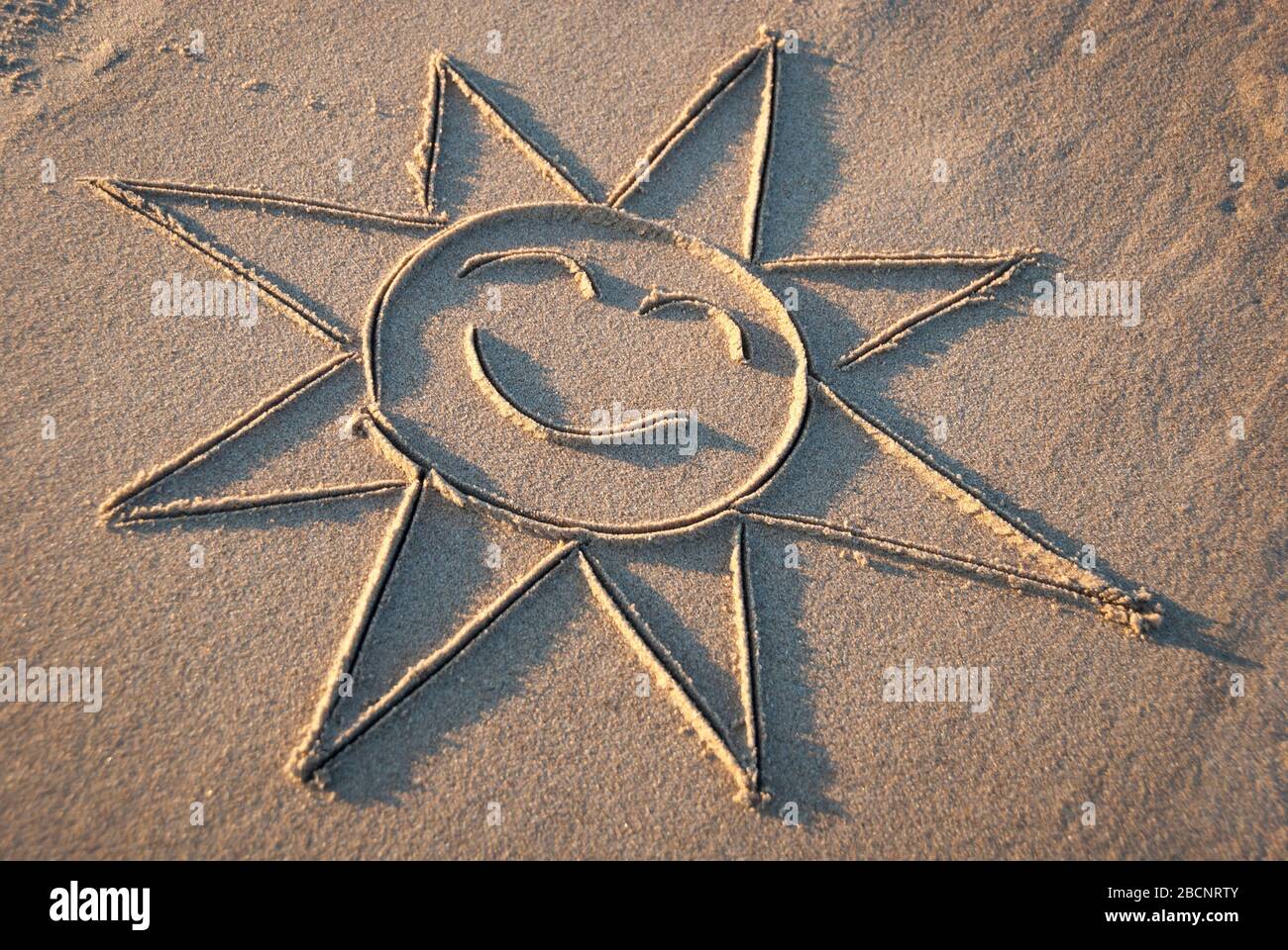 Fröhliche Sonne mit klassischen Strahlen, die mit einem Lächeln in glattem Sand an einem sonnigen Strand gezeichnet werden Stockfoto