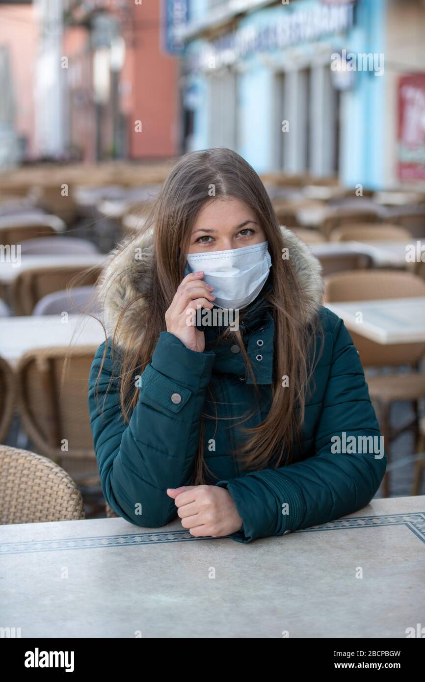 Ernste junge Frau mit schützender Gesichtsmaske, die draußen auf der leeren Café-Terrasse sitzt und während des coro mit jemandem auf ihrem Handy spricht Stockfoto