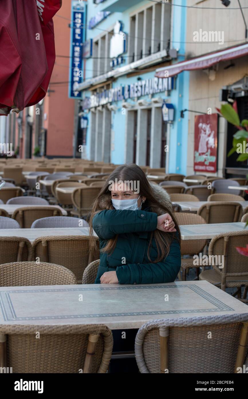 Ernste junge Frau mit schützender Gesichtsmaske, die draußen auf der leeren Café-Terrasse sitzt und während des Ausbruchs des Coronavirus hustet Stockfoto