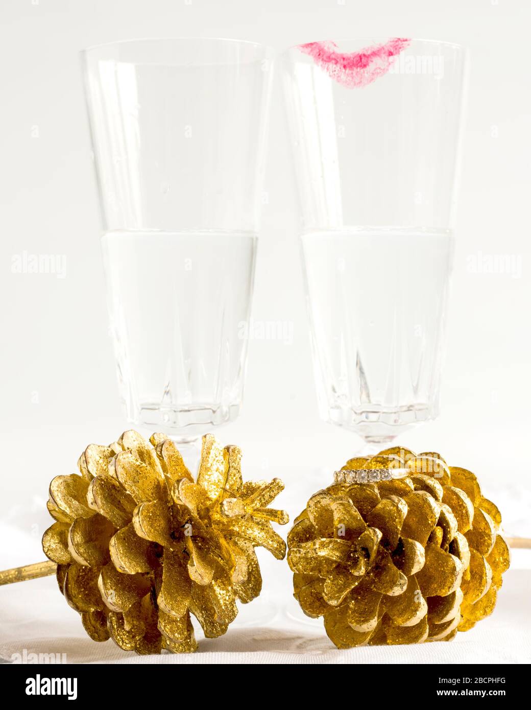 Zwei Champagnergläser, eine mit Lippenstift, hinter zwei goldenen Farnkegeln. Diamantring auf einem Kegel platziert. Wir feiern Engagement, standesamtliche Trauung, Hochzeit. Stockfoto