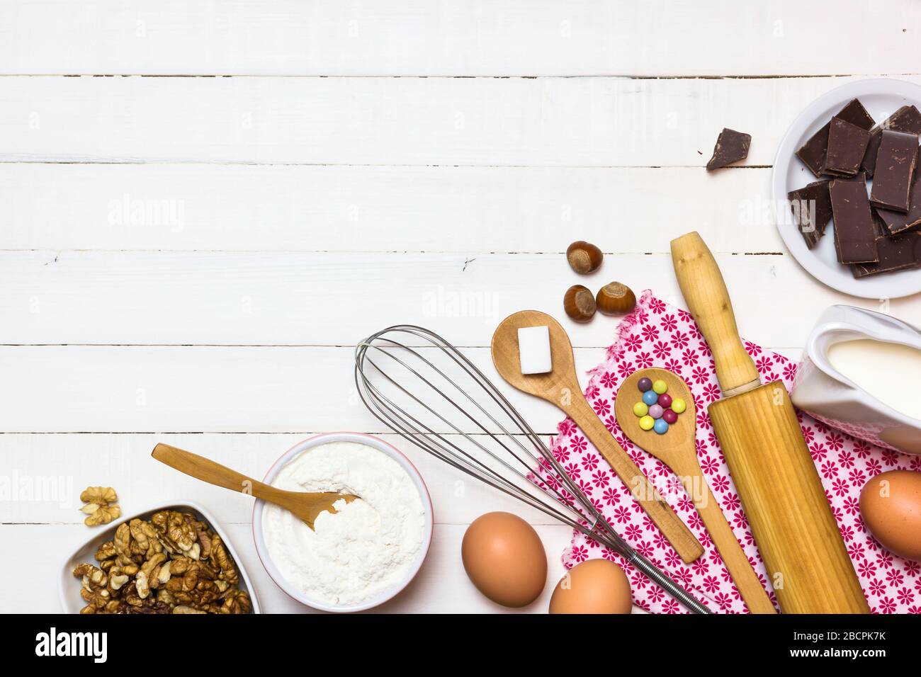 Zutaten und Utensilien zum Backen von Kuchen. Eier, Mehl, Milch, Zucker, Nüsse und Schokolade auf weißem Holztisch. Backen- oder Kochkonzept. Stockfoto