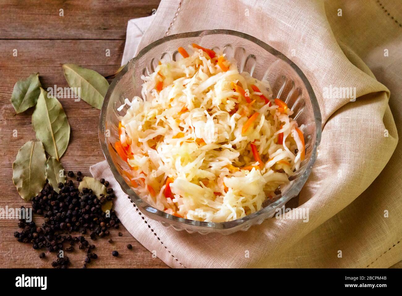 Hausgemachtes fermentiertes Sauerkraut in Glasschale auf rustikalem Holzhintergrund, LeinenServiette, Paprika, Lorbeerblätter Stockfoto