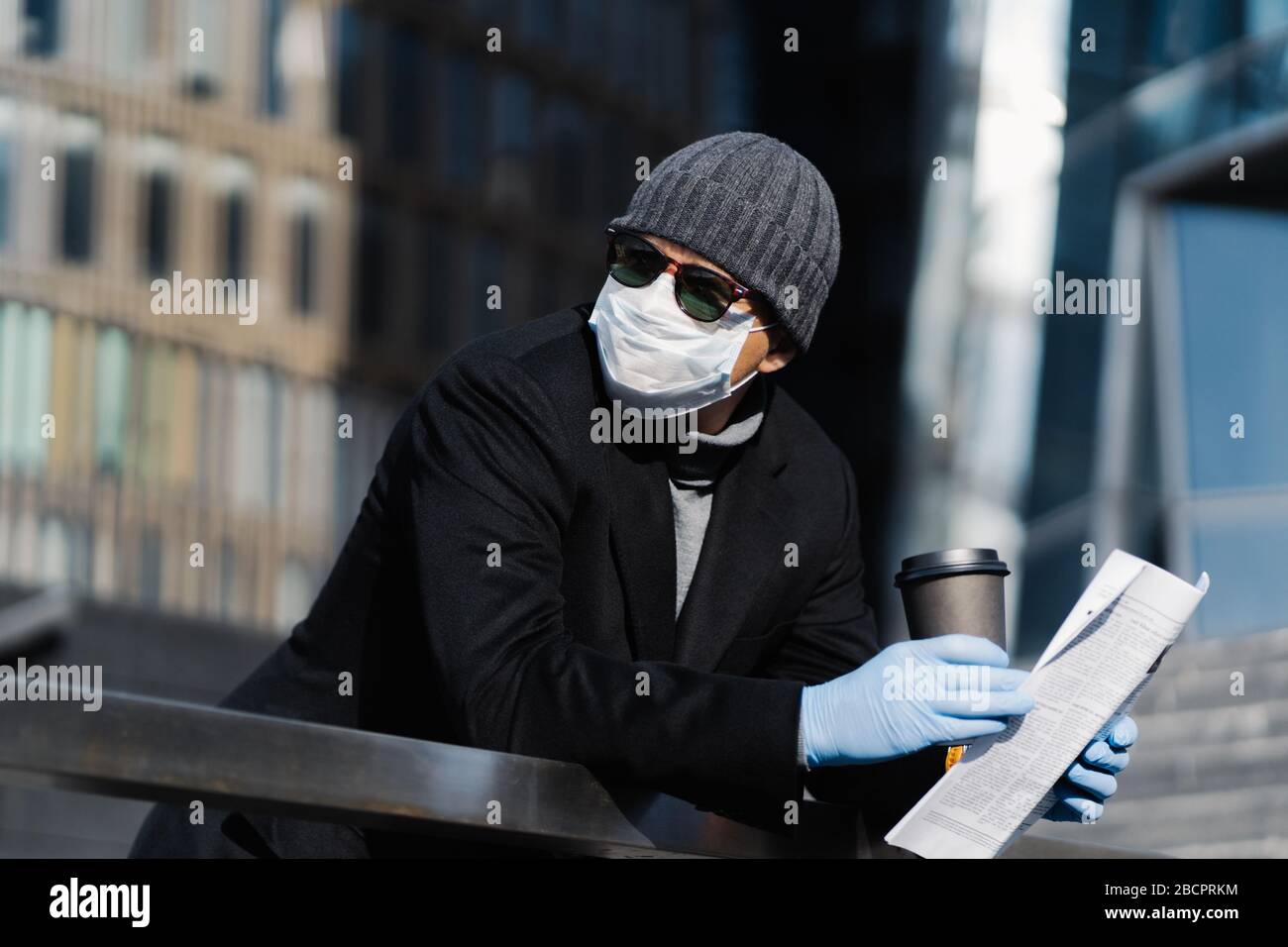 Junger Mann auf der Straße mit Kaffee und Zeitung zum Mitnehmen, trägt schützende medizinische Maske und Handschuhe, verhindert die Verbreitung von Coronavirus, sieht nachdenklich asid Stockfoto