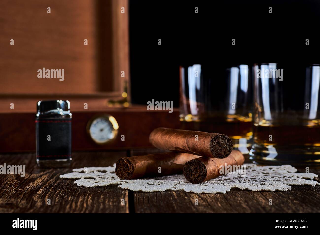 Stillleben mit drei kubanischen Zigarren, zwei Gläsern Whisky oder Rum, leichtere und Holzkiste mit Hygrometer. Alte Holztischplatte und schwarzer Hintergrund. Stockfoto