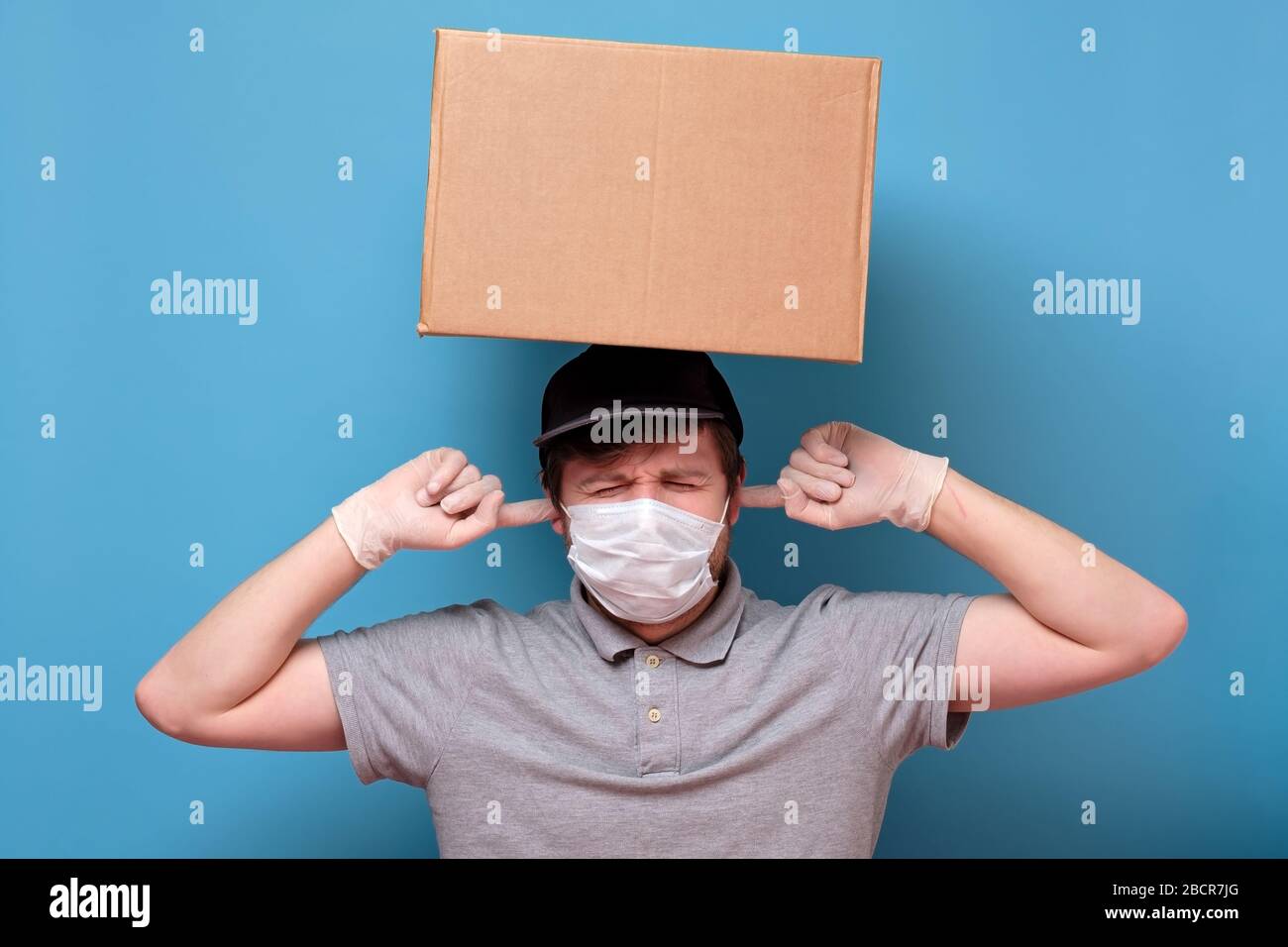 Kaukasischer junger Mann in medizinischer Maske, der ein Paket hält, das die Ohren vor Lärm bedeckt. Kein ansteckendes Lieferkonzept. Studio an blauer Wand gedreht. Stockfoto