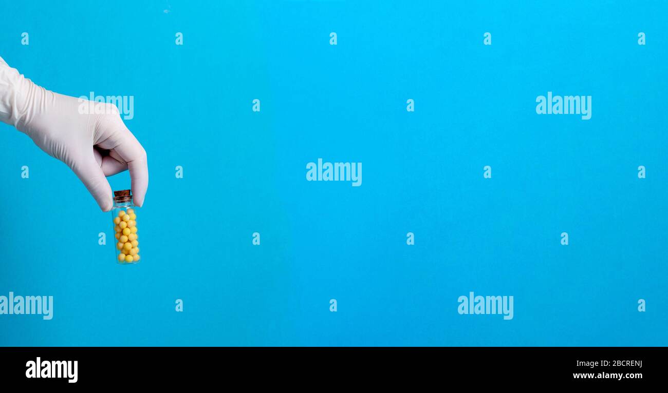Gelbe Globuli Glasflasche in Hand mit Handschuhen auf blauem Hintergrund. Banner. Kopierbereich. Stockfoto