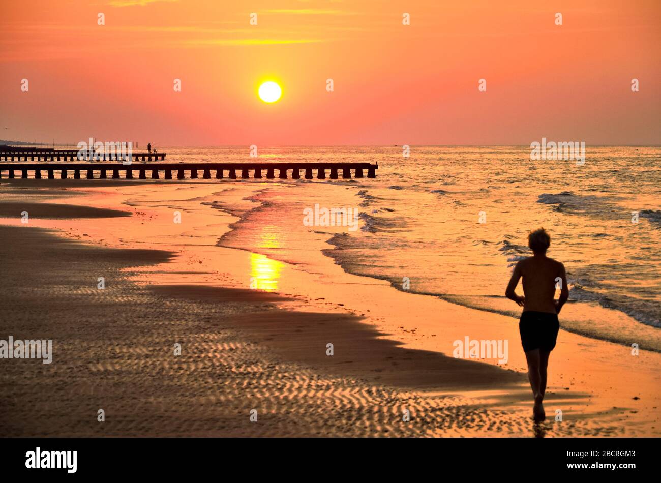 Silhouette des jungen Mannes, der bei Sonnenaufgang auf Strandsand läuft. Originaltapete aus dem aktiven Sommerurlaub. Stockfoto