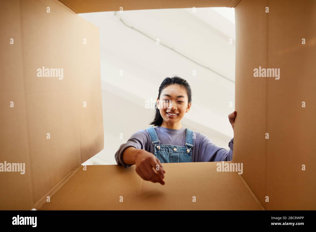 Der niedrige Blickwinkel beim Lächeln der asiatischen Frau, die beim Verpacken oder Auspacken in den Karton greift, um ein neues Zuhause zu finden, Platz für Kopien Stockfoto