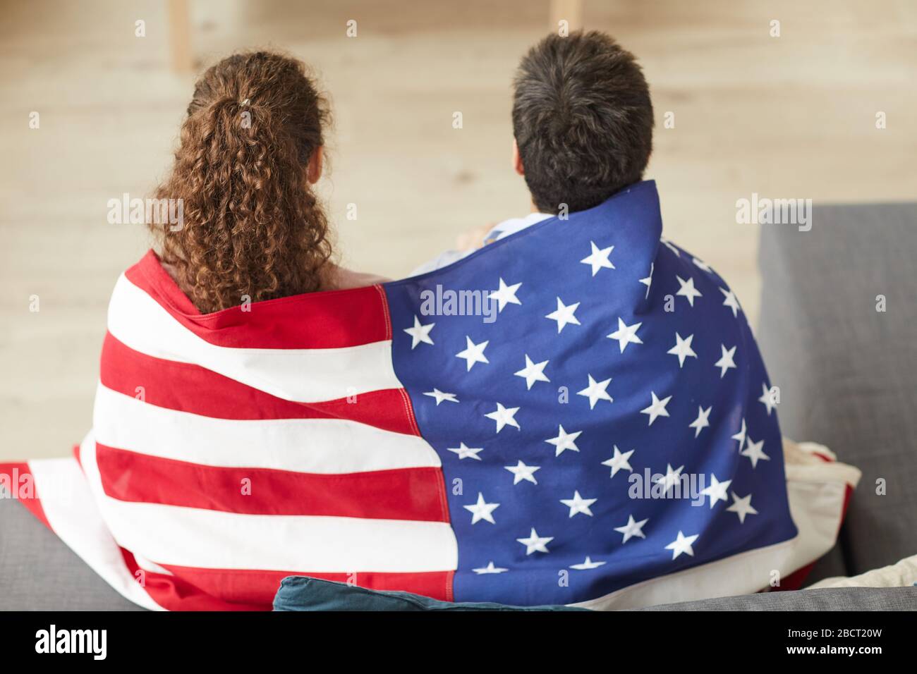 Rückansicht Portrait des jungen patriotischen Paares, das amerikanische Flagge hält, während man zu Hause Fernsehen kann, Kopierraum Stockfoto