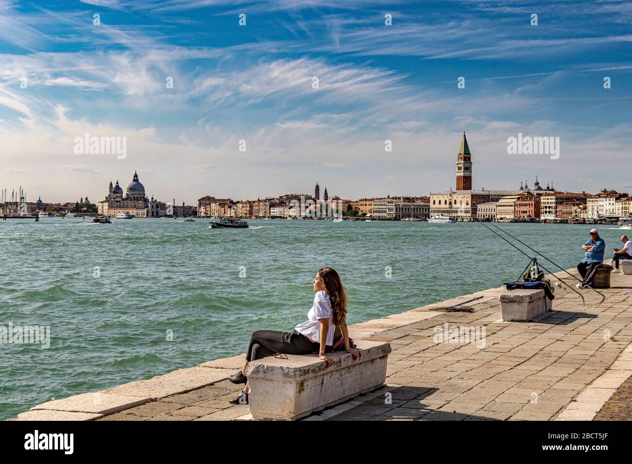 Frau auf einer Steinbank in der Sonne am Wasser in der Nähe von Arsenale mit St. Mark's Campanile und dem Dogenpalast im Hintergrund, Venedig Stockfoto
