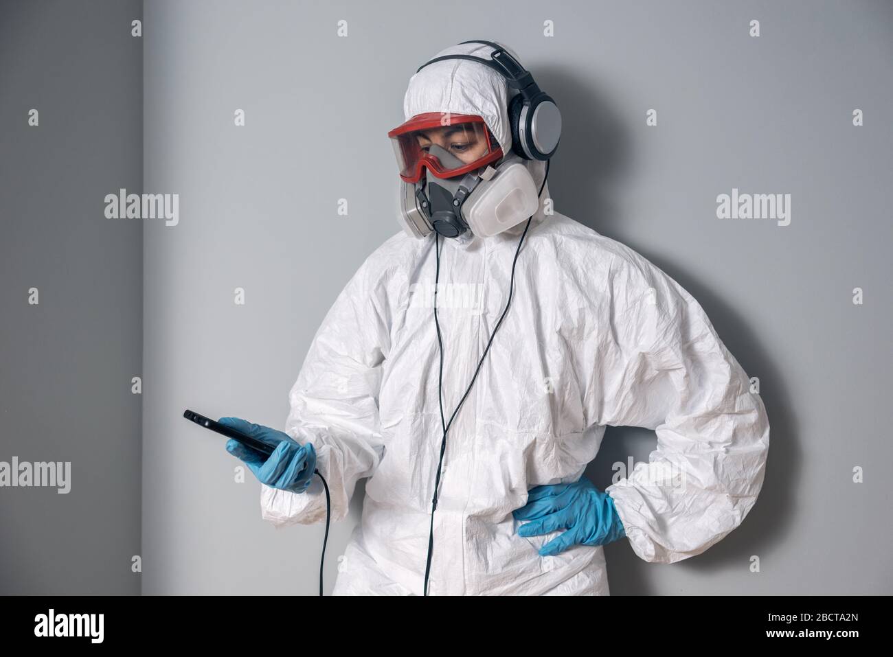 Frau mit Laborkittel, Nitrilhandschuhen, Schutzbrille, Gesichtsmaske und NBC-Schutzanzug, Blick auf das Smartphone-Handy und trägt musikalische Luftröhre Stockfoto