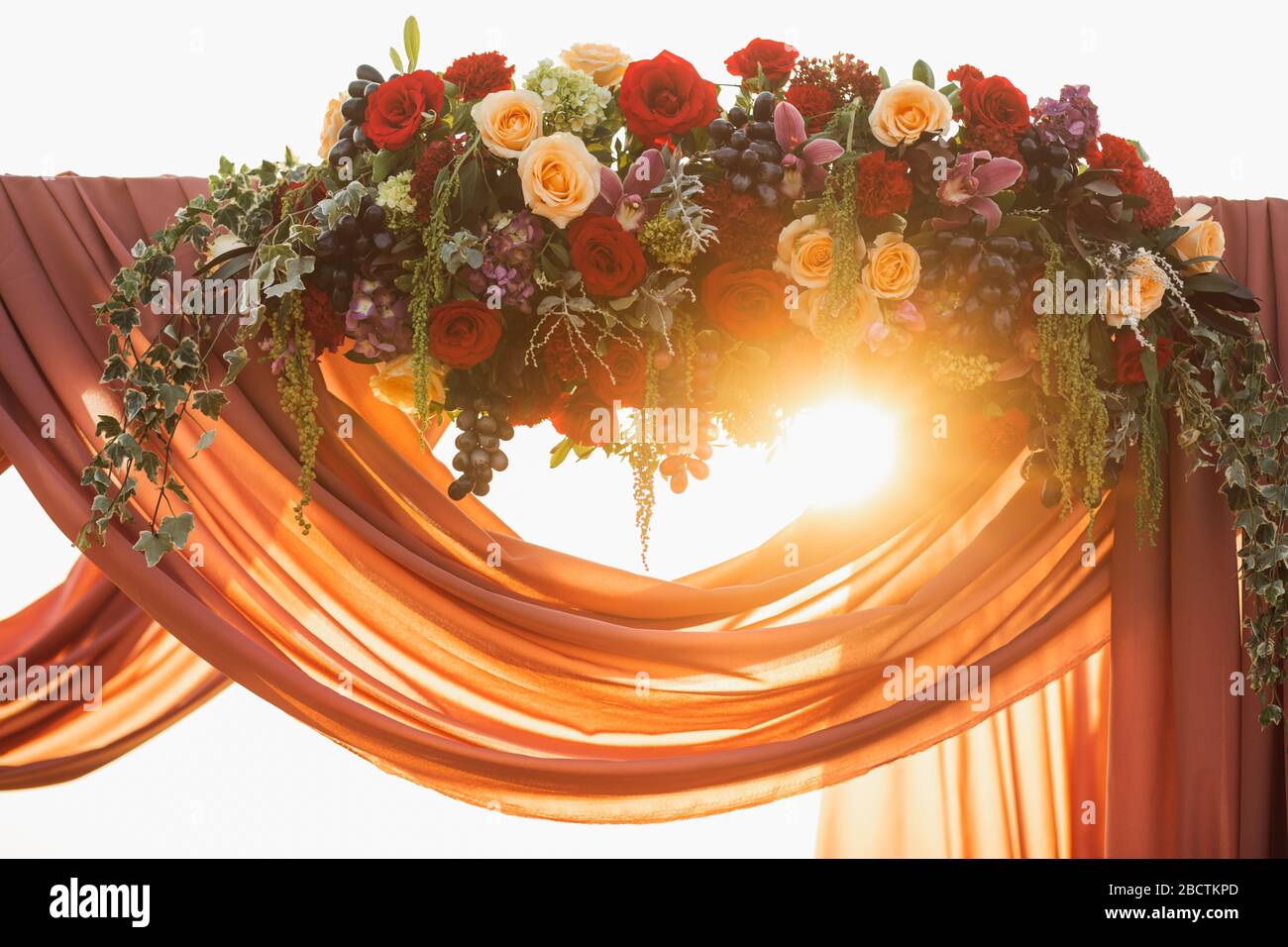 Dekoration des Hochzeitsbogens aus Holz bei der Zeremonie zum Sonnenuntergang im Freien. Rote und beige Rosenblüten, Trauben und Hängetuch. Wundervolle warme Sonne. Stockfoto