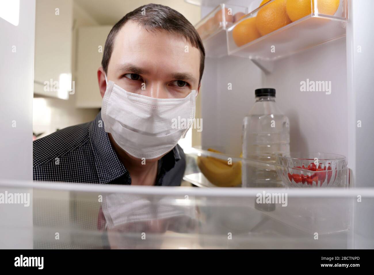 Der Mann in einer medizinischen Maske schaut mit Essen in den Kühlschrank. Ernährungskonzept, Fettleibigkeit während der Hausquarantäne bei einer Kovid-19-Coronavirus-Pandemie Stockfoto