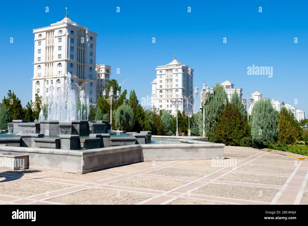 Platz in Ashgabat, Turkmenistan, mit Wasserbrunnen und mehreren neu errichteten weißen Marmorwohngebäuden dahinter. Stockfoto