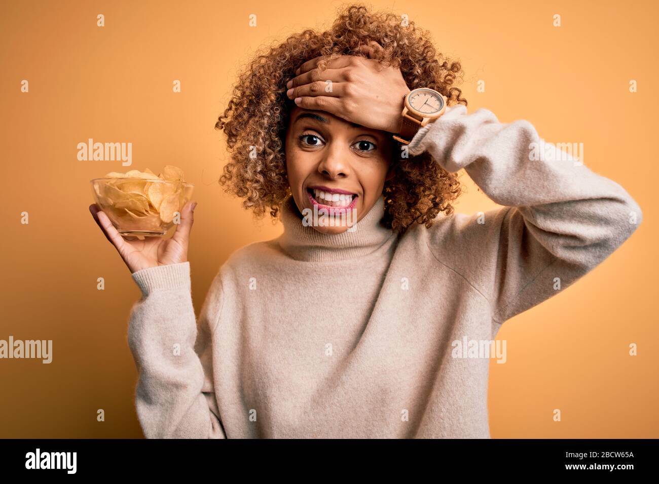 Junge afroamerikanische Curly Woman, die Schüssel mit Pommes Kartoffeln über gelbem Hintergrund mit Hand auf dem Kopf hält, schockiert mit Scham und Überraschung Stockfoto